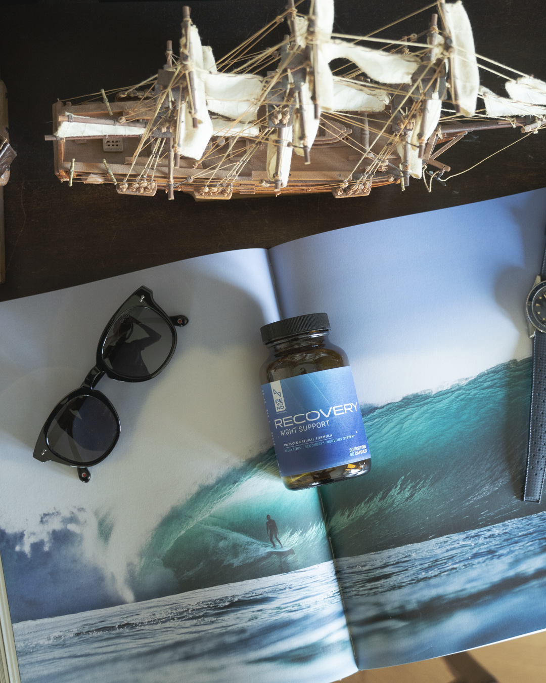 Model ship, sunglasses, and 'Recovery' supplement bottle on a magazine with a beach scene.