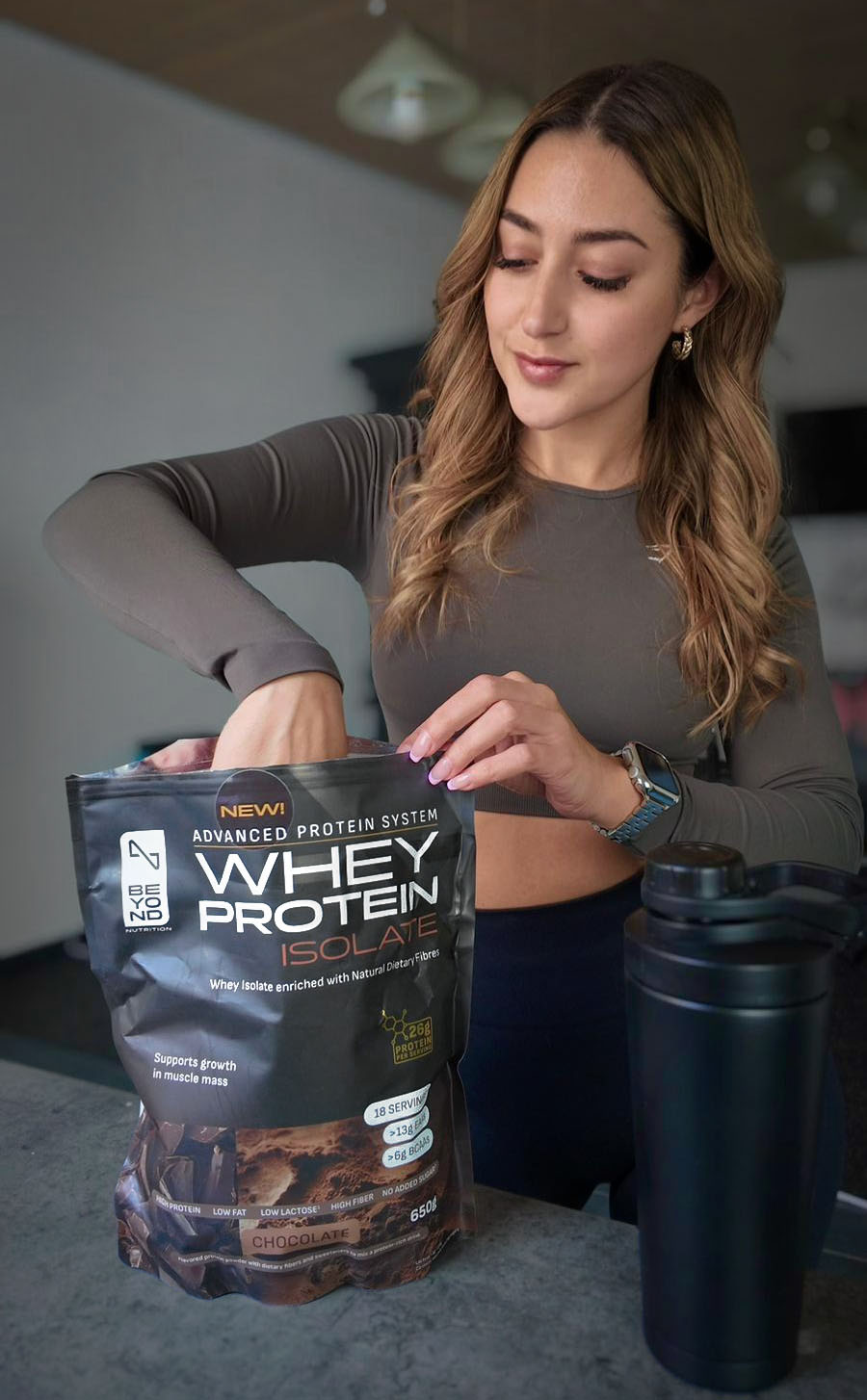 Woman holding a package of whey protein isolate from Beyond Nutrition with a black shaker bottle on a table.