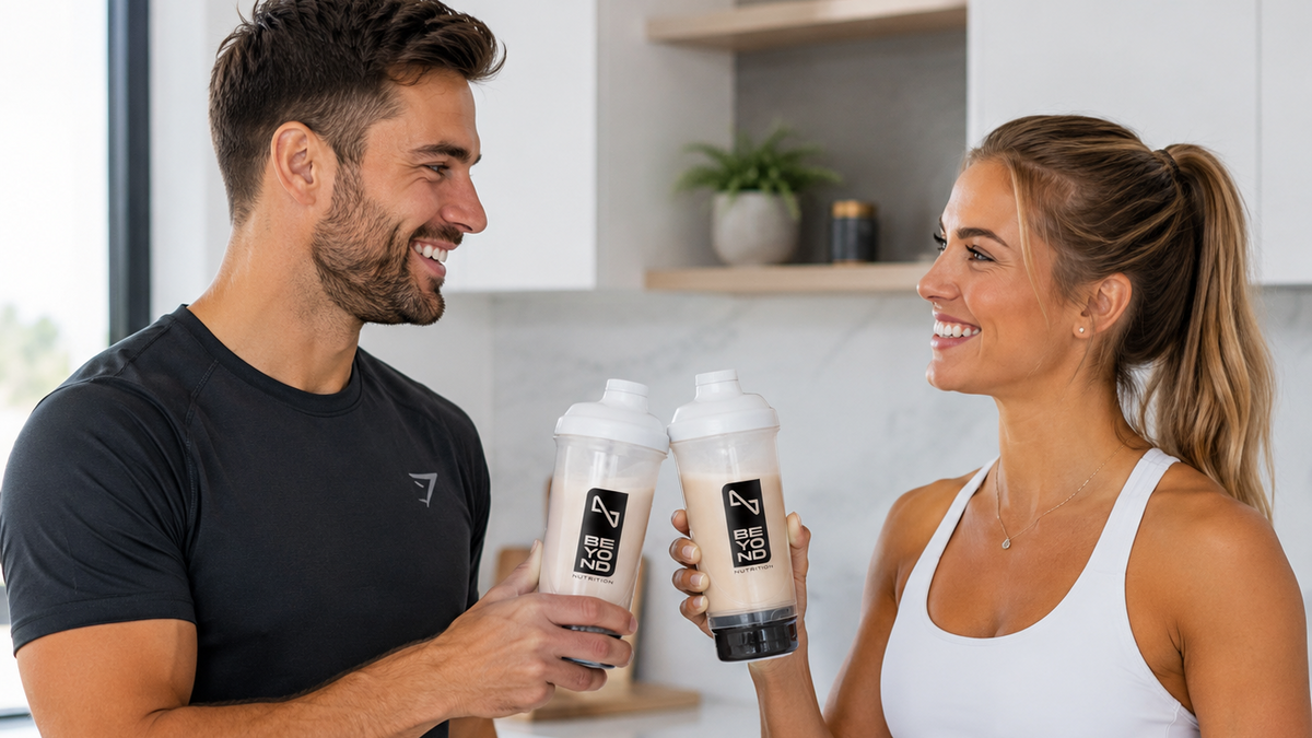 A man and a woman in athletic wear smile while holding two Beyond Nutrition shaker bottles with the same black-and-white logo. The man wears a dark gray t-shirt, and the woman wears a white tank top.