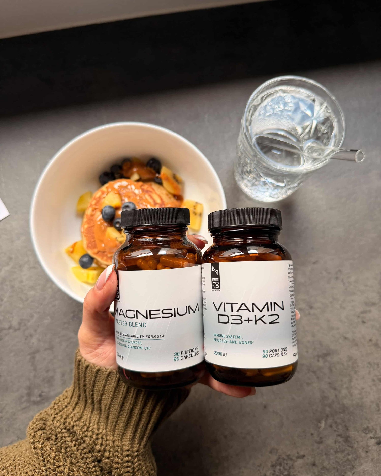 Two supplement bottles labeled 'Magnesium' and 'Vitamin D3+K2' held by a hand, with a bowl of food and a glass of water in the background.