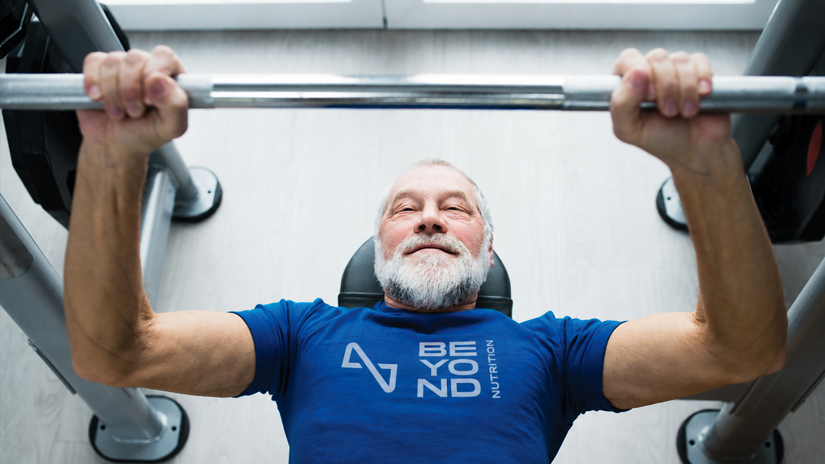 A man with a gray beard and mustache lies on a weight bench in a gym, lifting a barbell overhead. He is wearing a blue t-shirt with a white graphic that reads 'BEYOND NUTRITION'.