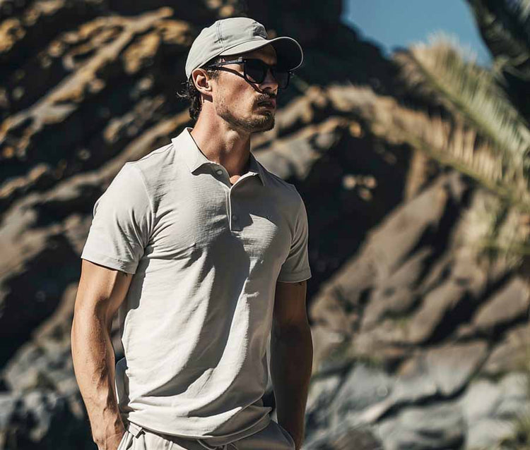 Man wearing a light gray polo shirt and cap standing against a rocky background