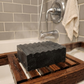 Black soap bar on a wooden tray in a bathroom setting