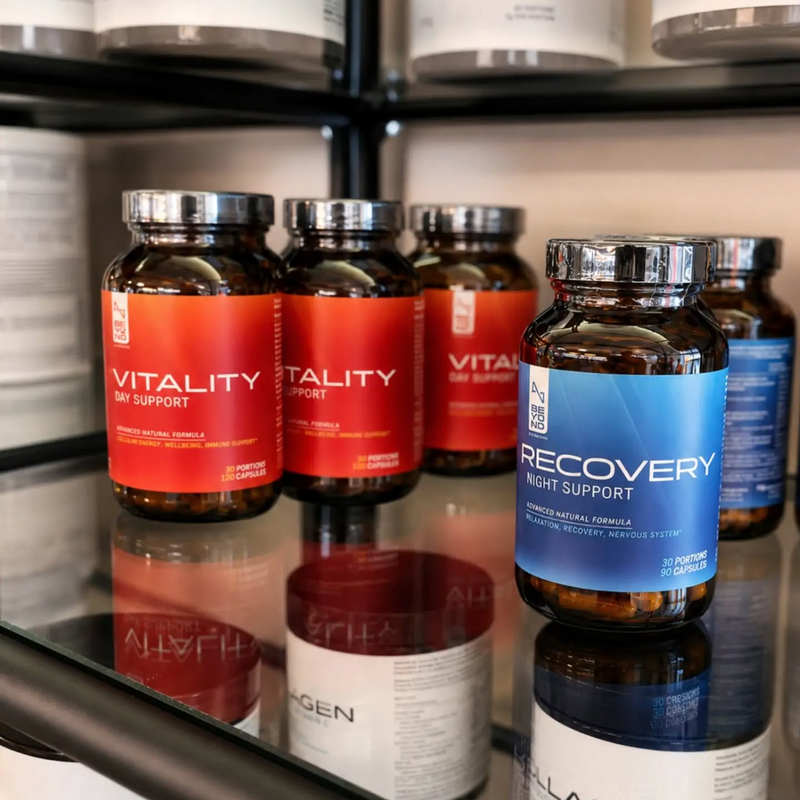 Several supplement bottles displayed on a glass shelf in a retail or warehouse setting. The most prominent product in the foreground is a dark blue bottle labeled 'Recovery Night Support' by Pure Encapsulations, with other red and orange bottles behind it labeled 'Vitality' by Life Extension.