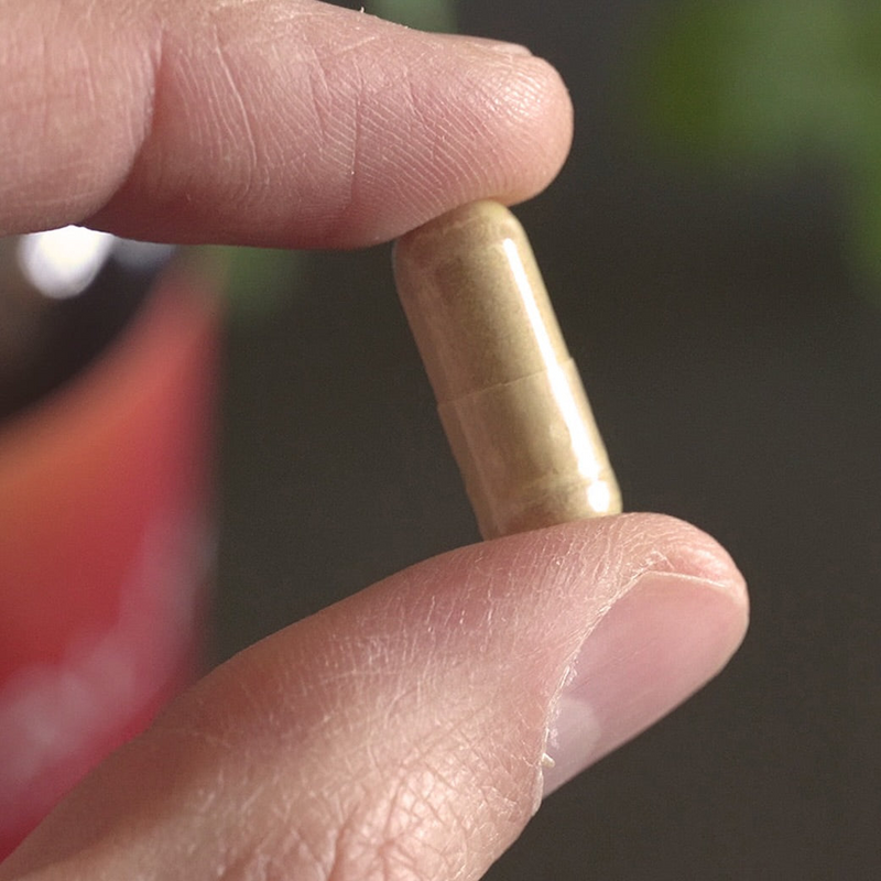 A close-up of a hand holding a small beige capsule pill between two fingers against a dark, blurred background.